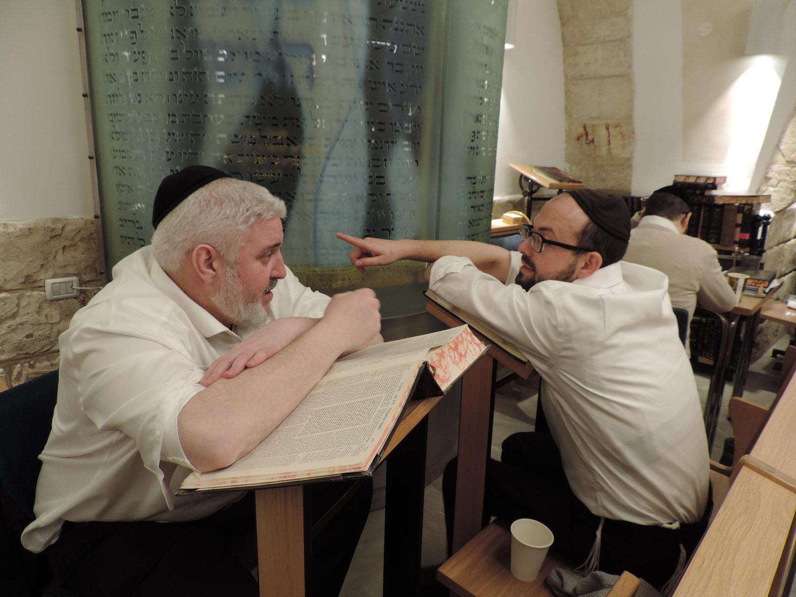 Two men arguing a sugya across their shtenders in the beis medrash, one pointing to make his case
