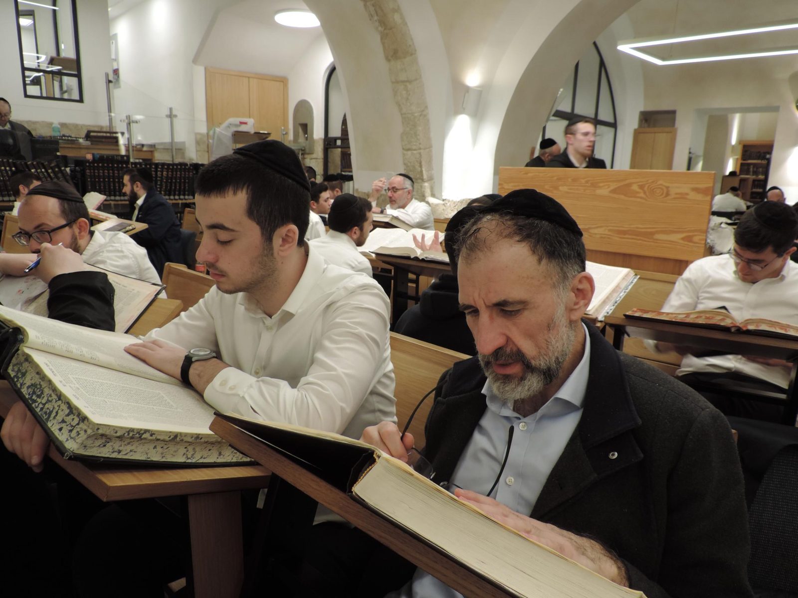 A young man and an older man side by side at their shtenders, each following his own open Gemara, the beis medrash busy around them