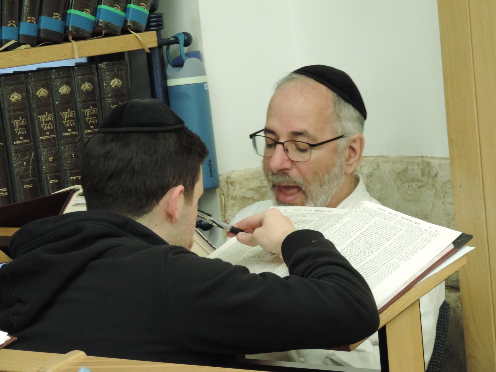 An older man and a young talmid learning together from one open Gemara, the older man mid-explanation