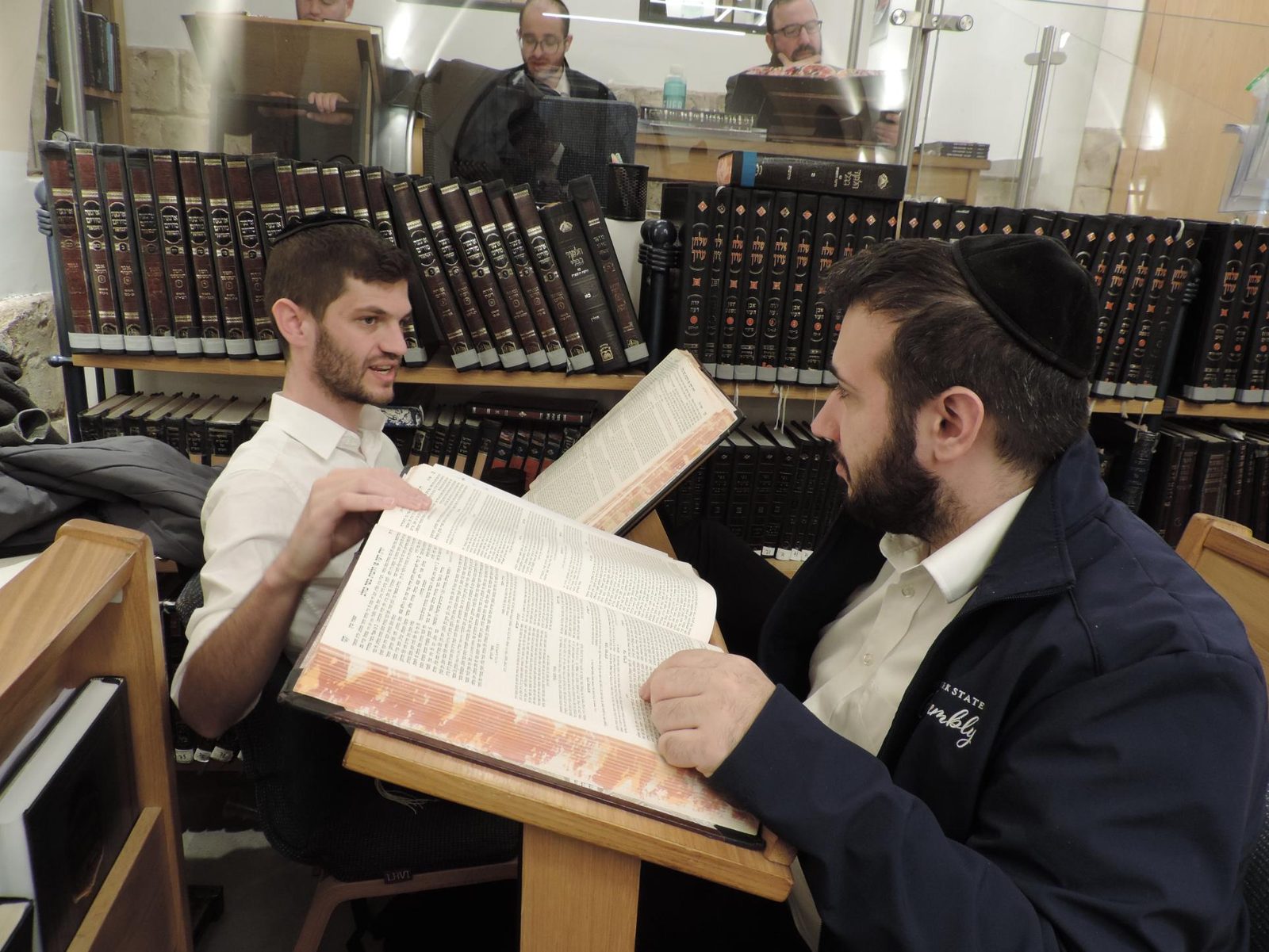A chavrusa pair mid-sugya, hands moving over an open Gemara, shelves of seforim behind them